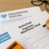 Mano rellenando formulario donde s indican las obligaciones del Ingreso Mínimo Vital 2025, con documentos de Seguridad Social, declaración de la renta y calculadora en escritorio.